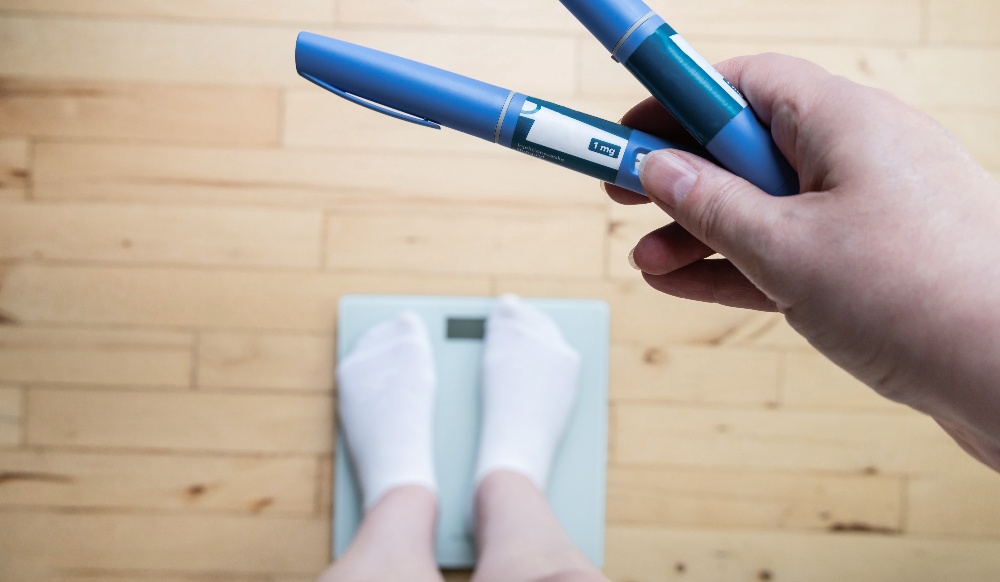 A patient holds a GLP-1 injector in hand while standing on a scale. 